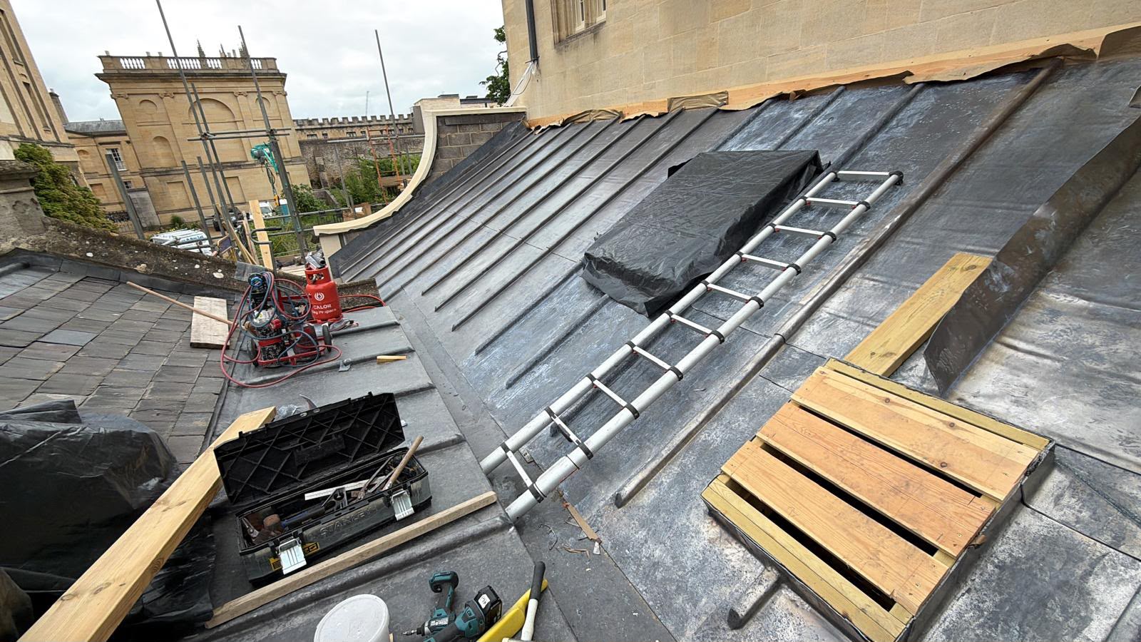 The new Deanery extension roof part way through leadwork, with finished lead bays on one side, underlay and timber still exposed on the other, and roofers’ tools and a ladder on the slope.