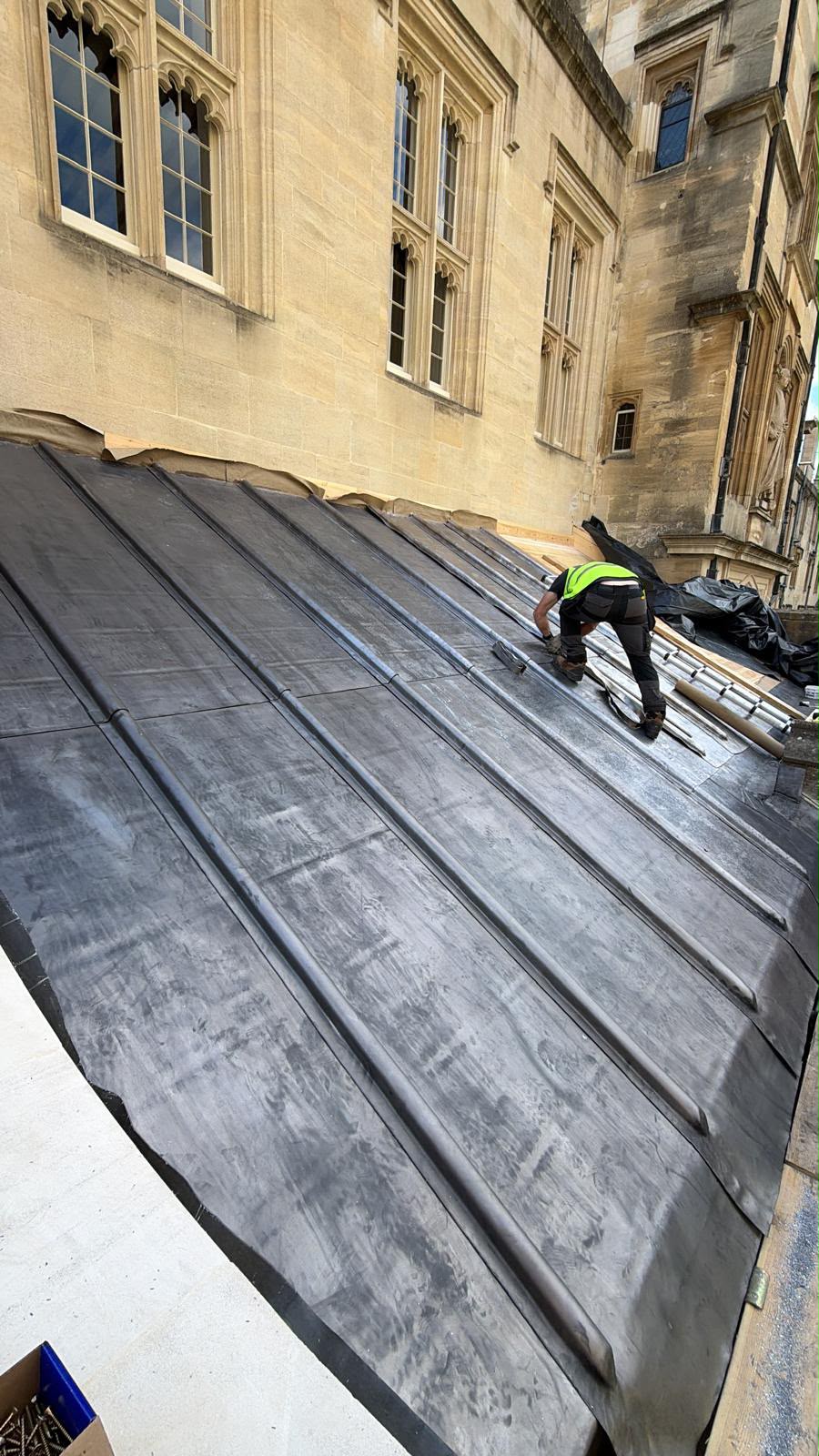 A roofer in a hi-vis vest kneeling on the new lead roof, working lead by hand along a wood-cored roll beneath the Deanery’s traceried stone windows.