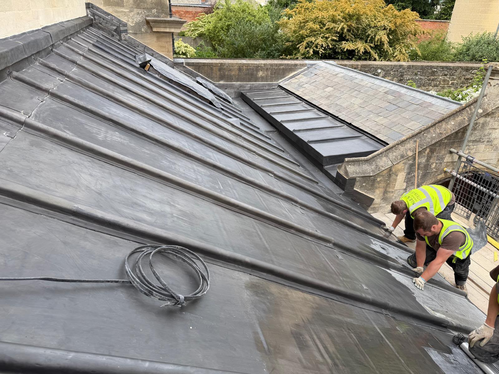 Two roofers dressing a fresh sheet of milled lead into place on the Deanery extension roof, with completed lead bays and a neighbouring slate roof beyond.